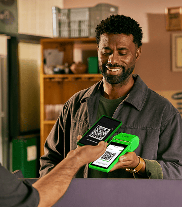 Empreendedor segurando uma maquininha de cartão Stone enquanto alguém paga um QR Code pelo celular