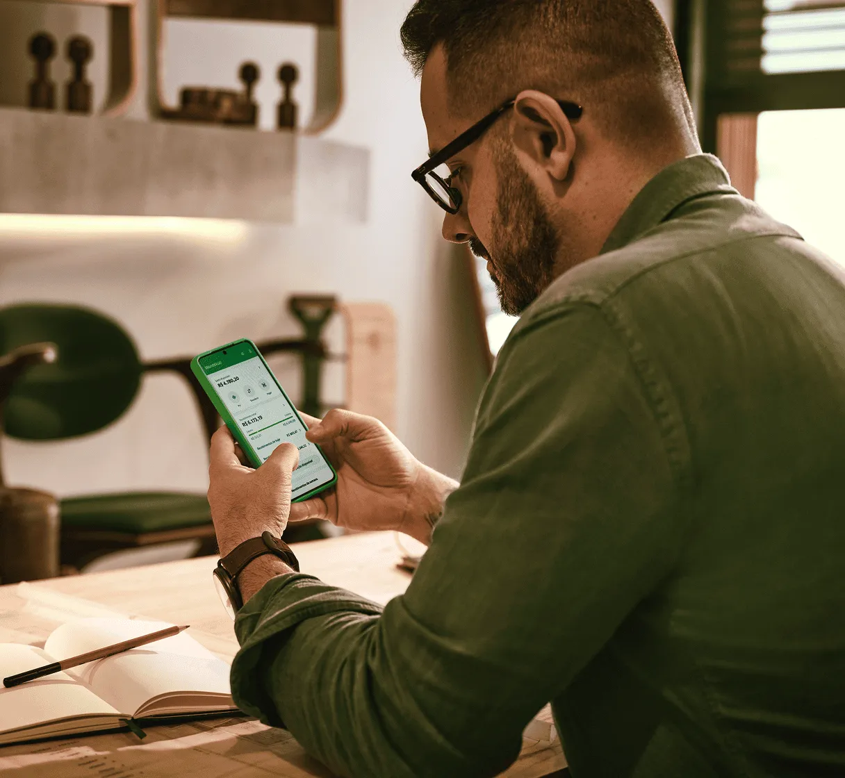 Um homem de óculos e barba, visto de costas, concentra-se no celular que segura. A tela do celular exibe o aplicativo Stone. Ele está em uma mesa de madeira com um caderno aberto e lápis.