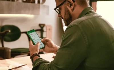 Um homem de óculos e barba, visto de costas, concentra-se no celular que segura. A tela do celular exibe o aplicativo Stone. Ele está em uma mesa de madeira com um caderno aberto e lápis.