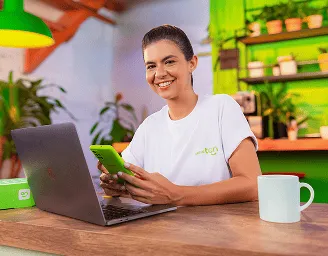 Consultora do Renda Extra Ton sentada numa cadeira e sorrindo olhando o celular e computador em uma sala decorada com elementos na cor verde.